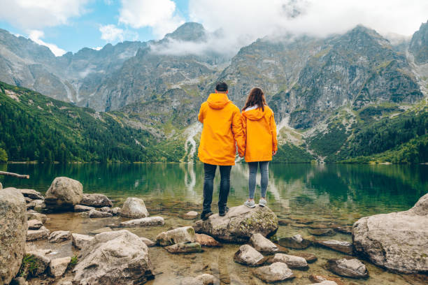 homme avec la femme dans le imperméable jaune au jour ensoleillé d’automne regardant le lac dans les montagnes de tatra - monts de tatra photos et images de collection