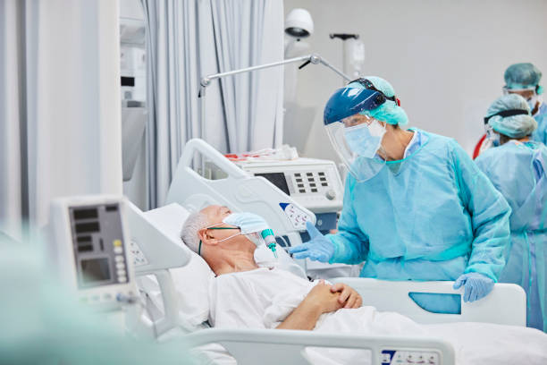 Doctor Talking to Senior Patient In Hospital During COVID-19 Doctor talking to patient in hospital ward. Healthcare professionals are in blue coveralls. They are treating senior male for coronavirus. medical ventilator photos stock pictures, royalty-free photos & images
