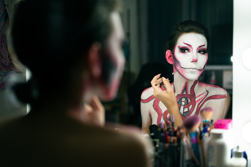 Reflection portrait of young woman in front of a mirror. She is painting her face and body for the Halloween.