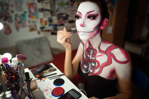 Close up view of  a young woman artist applying make-up for the Halloween. Stitches on her cheeks and red snakes over upper part of her body.