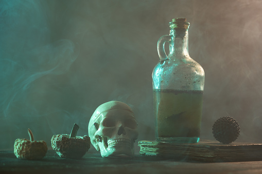 Three rotten pumpkins, a black antique book, virus shaped object and a bottle full of dirty poisonous liquid on wooden desk for halloween concept. The background is foggy and black. Shot with a full frame mirrorless camera.