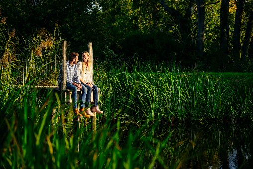 Couple in early 20s sitting side by side and talking in late afternoon sunshine at end of small pier overlooking rural pond surrounded by tall grassy reeds.