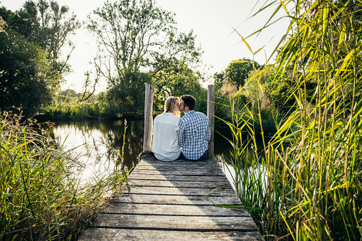 Rear view of Caucasian couple in early 20s wearing casual clothing and kissing as they sit side by side at end of rural pier in late afternoon sunshine.