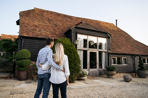Rear view of British couple wearing casual clothing and standing with arms around each other admiring their home converted from traditional wooden barn.