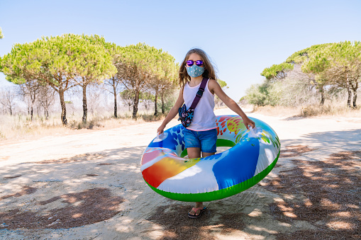 girl with a mask on her face and glasses walking with a colorful inflatable mattress wheel with her hands to enjoy the vacations in the middle of the coronavirus pandemic covid 19
