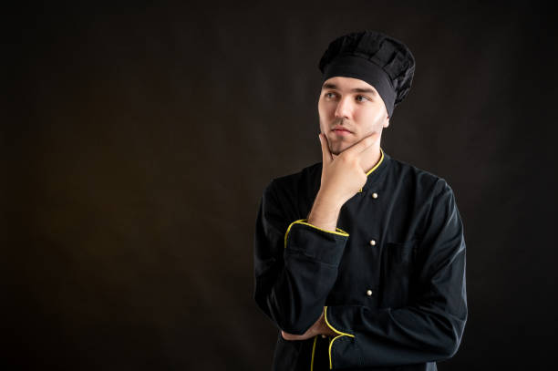istock Young male dressed in a black chef suit holding hand under his chin, having doubtful 1277109295