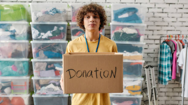 istock Caucasian guy holding donation box and looking at camera, posing in front of rack and boxes full of clothes, Young volunteer working for a charity 1277113672
