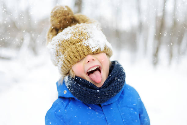 Funny little boy in blue winter clothes walks during a snowfall. Outdoors winter activities for kids. Funny little boy in blue winter clothes walks during a snowfall. Outdoors winter activities for kids. Cute child wearing a warm hat low over his eyes catching snowflakes with his tongue eye catching stock pictures, royalty-free photos & images