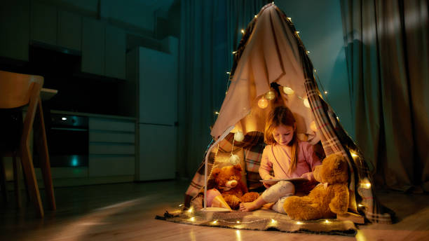 uma garotinha bonita usando pijamas sentado em um chão descalço em uma cabana feita de xadrez jogando em seu tablet sorrindo e tendo ursinhos e guirlandas com ela ao redor - teepee - fotografias e filmes do acervo