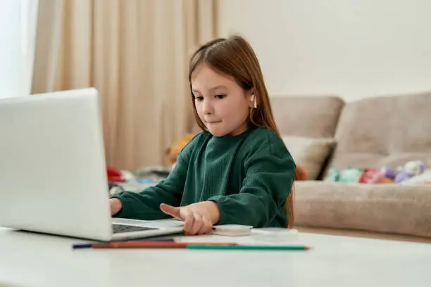 A cute girl decided to make her distance education hometask and moving a laptop on a table while sittining alone in a big bright guestroom wearing a green sweater and wireless earphones. Homeschooling