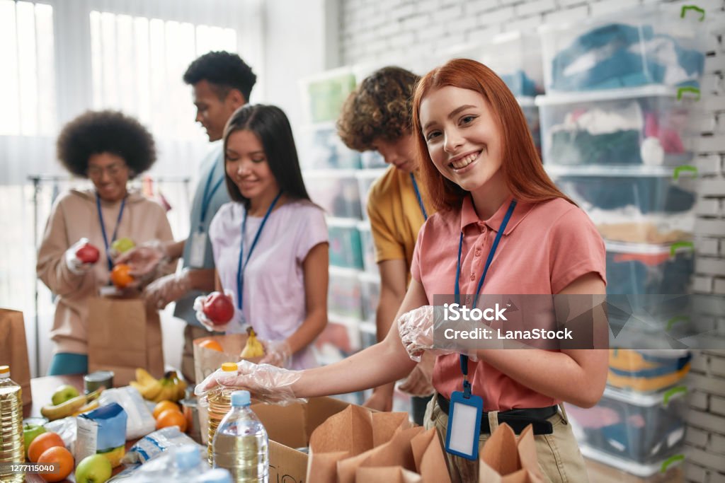 Young female volunteer smiling at camera while packing food and drinks donation into paper bags and box, Small group of people working in charitable foundation Young female volunteer smiling at camera while packing food and drinks donation into paper bags, Small group of people working in charitable foundation, Selective focus on girl, Horizontal shot Volunteer Stock Photo