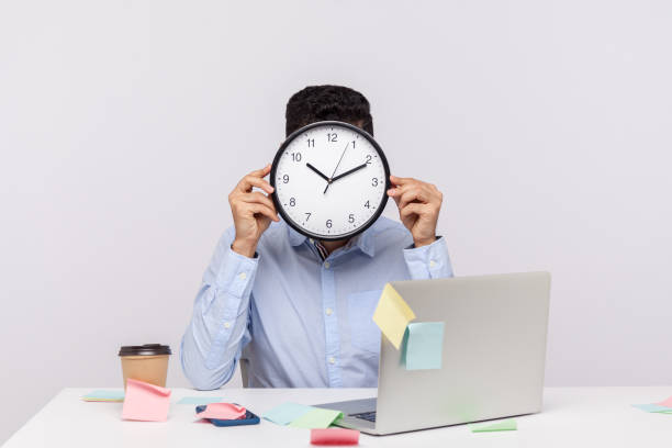 istock Overtime work. Anonymous man employee sitting in office workplace, hiding face behind clock, sticky notes 1277118406
