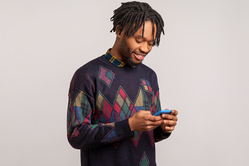 Cheerful satisfied african man with dreadlocks in casual style sweatshirt chatting smartphone with toothy smile on face, happy with 4g connection. Indoor studio shot isolated on gray background