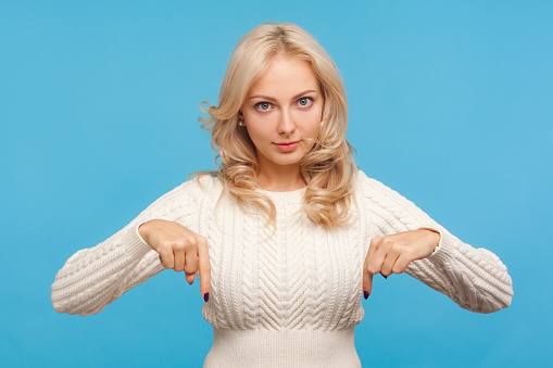 Confident bossy blond woman in white knitted sweater pointing fingers down, asking to act here and now immediately. Indoor studio shot isolated on blue background