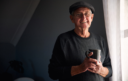 Portrait Of Senior Man Wearing Cap Relaxing Standing By Window At Home With Hot Drink