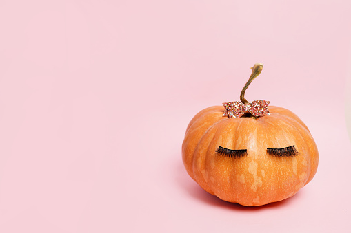 Pumpkin with false eyelashes and bow on pink background. Autumn, fall, halloween concept. copy space