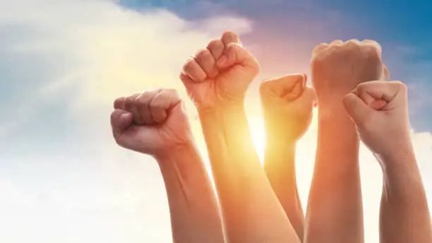Rising fist of adult people, male and female, over dramatic blue sky with sun light, anger protest revolution inspiration motivation or teamwork concept