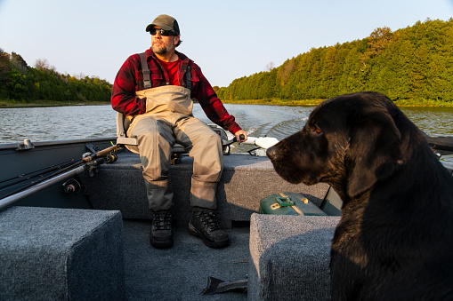 A mature male fisherman in a fishing boat with his labrador retriever dog on his lap.