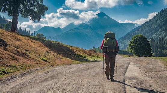 Tourist with backpack and trekking poles on road at mountains background at sunny day