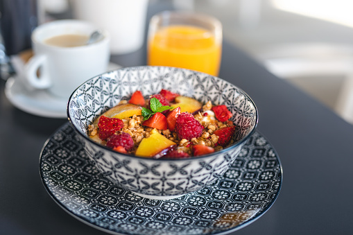 Healthy breakfast: Bowl with cereals, greek yogurt and fruits (strawberries, raspberries and nectarine) decorated with mint leaf. Defocused cup of coffee and glass of orange juice in the background.