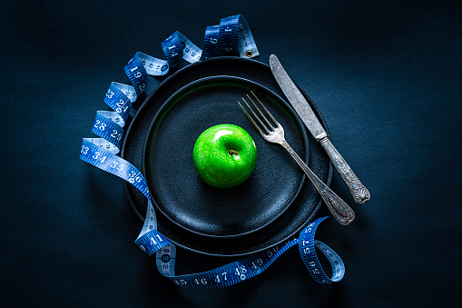Healthy eating concept: Top view of a black plate with a green apple on it shot on black background. A blue tape measure is all around the plate. A fork and knife are on each side of the plate. Predominant colors are black and green. High resolution 42Mp studio digital capture taken with SONY A7rII and Zeiss Batis 40mm F2.0 CF lens