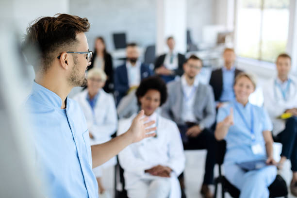 Male healthcare worker giving presentation to group of people in board room. Young doctor talking to group of his colleagues and business people during presentation in convention center. seminar stock pictures, royalty-free photos & images
