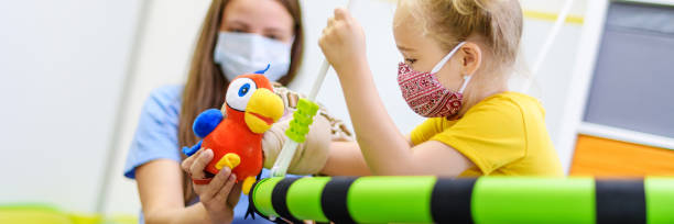Toddler girl in child occupational therapy session doing playful exercises with her therapist during Covid - 19 pandemic, both wearing protective face masks. Web banner. Toddler girl in child occupational therapy session doing playful exercises with her therapist during Covid - 19 pandemic, both wearing protective face masks. Web banner. occupational therapy photos stock pictures, royalty-free photos & images