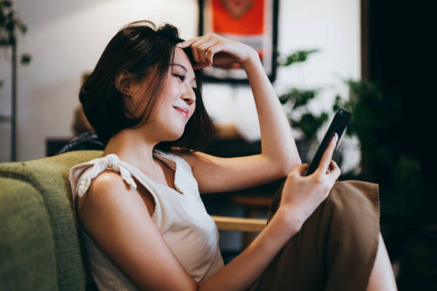 istock Beautiful smiling young Asian woman chilling at home, using smartphone while sitting on the sofa in the living room 1277156471