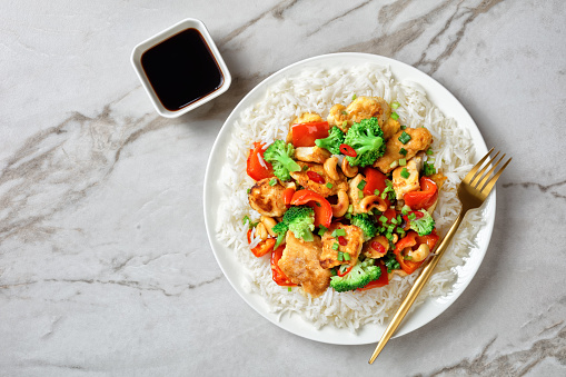 Asian cashew chicken with vegetables stir-fry: broccoli, bell pepper, hot red chili, and soya sauce on a white plate with fork  on a white marble background, top view, close-up