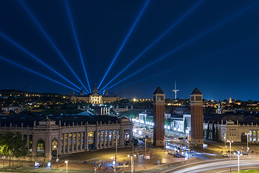 Panorama from the Plaza de Espana (Spain Square) to Montjuic and the National Art Museum of Catalonia with beams of searchlights cutting through the dark sky.