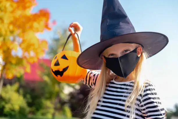 Photo of Halloween kids mask. Portrait blonde girl in witch costume with pumpkin bucket. Child wearing black face masks outdoors protecting from COVID-19