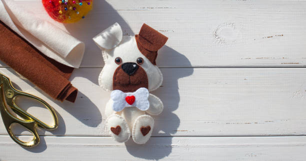 A beautiful felt puppy with a brown ear and muzzle, with a bone and a red  heart on a white boards background. Tailoring scissors, pins, pincushion. Creativity, hobbies, sewing toys with the child.