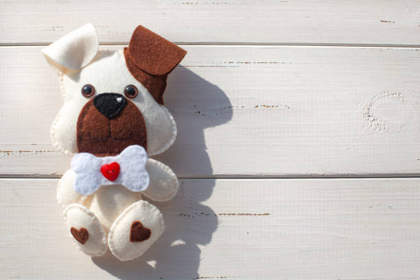 A beautiful white felt puppy with a brown ear and muzzle, with a bone and a red button in a heart on a white boards background. Creativity, hobbies, sewing toys. Needlework with the child. Copy space.