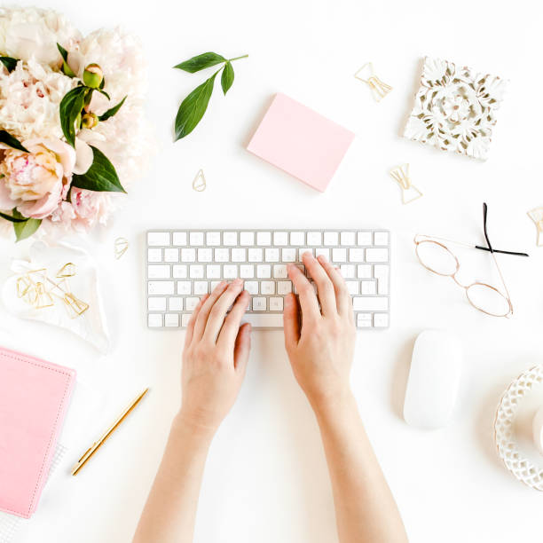 Flat lay women's office desk. Female workspace with female hands, computer, pink peonies bouquet, accessories on white background. Top view