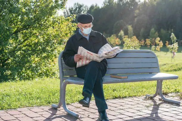 aged senior person wearing facemask sitting on wooden bench in park and reading newspaper new normal concept