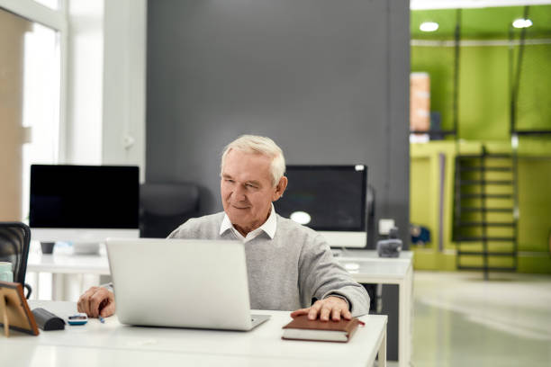 istock Portrait of cheerful aged man, senior intern looking focused at the screen while using laptop, sitting at desk, working in modern office 1277192412