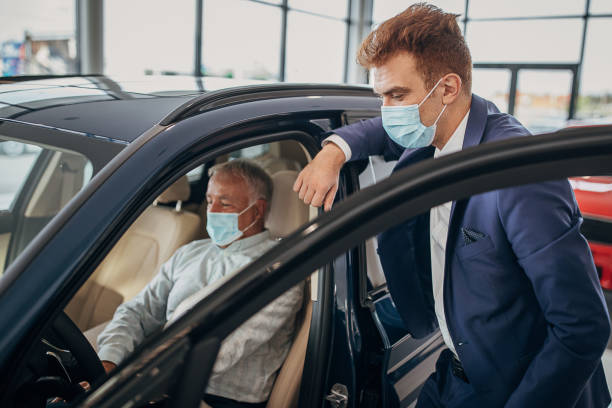 Car saleperson with protective face mask showing a car to senior male customer Car saleperson with protective face mask showing a car in a showroom to senior male customer who is also wearing protective face mask. car rental covid stock pictures, royalty-free photos & images