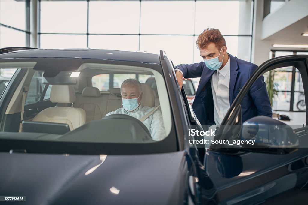 Young car saleperson with protective face mask showing a car to senior male customer Car saleperson with protective face mask showing a car in a showroom to senior male customer who is also wearing protective face mask. Protective Face Mask Stock Photo