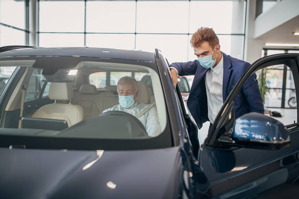 Young car saleperson with protective face mask showing a car to senior male customer Car saleperson with protective face mask showing a car in a showroom to senior male customer who is also wearing protective face mask. car ownership photos stock pictures, royalty-free photos & images