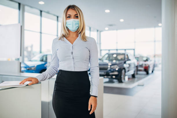 Portrait of young female car dealer with protective face mask in car showroom One young female car dealer with protective face mask working in car showroom. car rental covid stock pictures, royalty-free photos & images