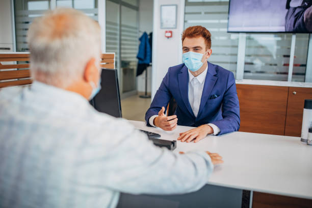 Male customer and young car saleperson with protective face mask talking about buying a new car Senior man and car saleperson with protective face masks sitting at the desk and talking about buying a new car. car rental covid stock pictures, royalty-free photos & images