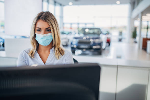 One female car dealer with protective face mask working on computer in car showroom One young female car dealer with protective face mask working on computer in car showroom. car rental covid stock pictures, royalty-free photos & images