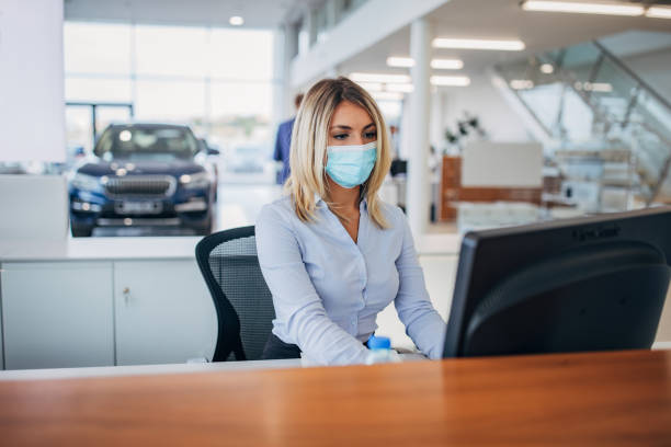 One female car dealer with protective face mask working on computer in showroom One young female car dealer with protective face mask working on computer in car showroom. car rental covid stock pictures, royalty-free photos & images