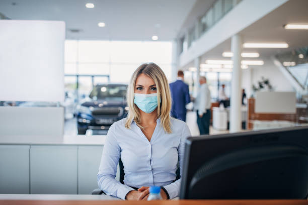 Female car dealer with protective face mask working in car showroom One young female car dealer with protective face mask working on computer in car showroom. car rental covid stock pictures, royalty-free photos & images