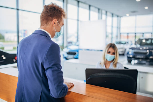Young saleswoman and male customer with protective face masks in car showroom Young saleswoman and young male customer with protective face masks talking about the possibility of buying a new car in car showroom. car rental covid stock pictures, royalty-free photos & images