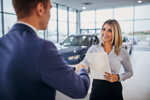 Young female car dealer shaking hands with young male customer after a successful car purchase in a car showroom. Focus on a female car dealer.