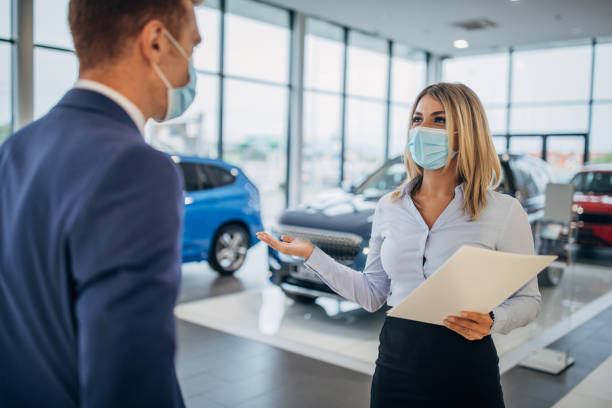 Female car dealer and male customer with protective face masks talking in a car showroom Young female car dealer and young male customer with protective face masks talking in a car showroom. car rental covid stock pictures, royalty-free photos & images