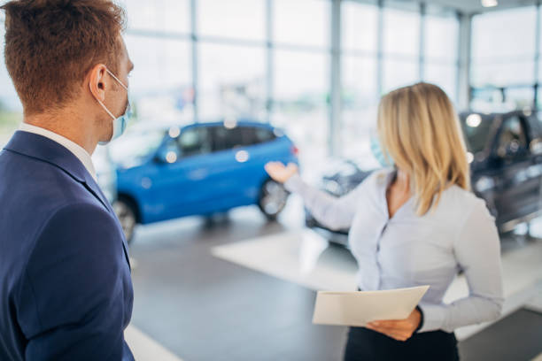Young car dealer and young man with protective face masks talking in a car showroom Young female car dealer and young male customer with protective face masks talking in a car showroom. car rental covid stock pictures, royalty-free photos & images