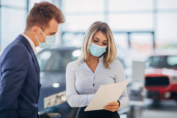 Young female car dealer and young man with protective face masks going through paperwork in a showroom Young female car dealer and young male customer with protective face masks talking about car purchase agreement in a car showroom. car rental covid stock pictures, royalty-free photos & images