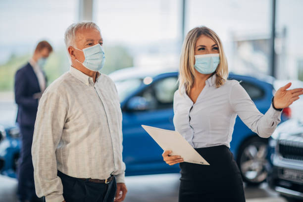 Young female car dealer and senior man with protective face masks talking in a car showroom Young female car dealer and senior male customer with protective face masks talking in a car showroom. car rental covid stock pictures, royalty-free photos & images